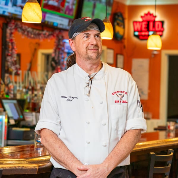 Chef Mike Moquin in a white chef coat with the Shortstop Bar & Grill logo, standing at a bar.