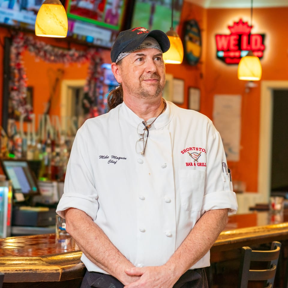 Chef Mike Moquin in a white chef coat with the Shortstop Bar & Grill logo, standing at a bar.