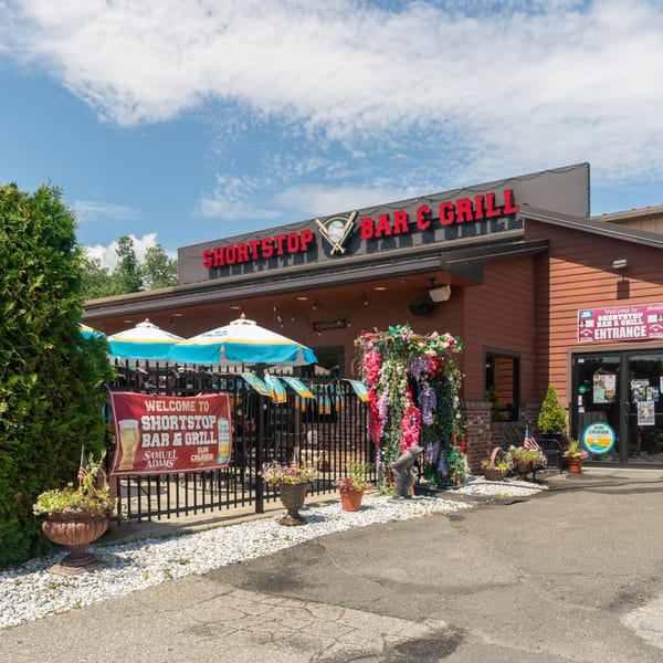 Exterior of Shortstop Bar & Grill with outdoor patio seating under umbrellas and a welcome banner.