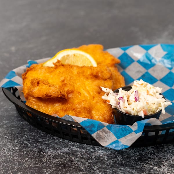 Golden fried fish with a lemon wedge and coleslaw in a black basket with blue checkered paper.
