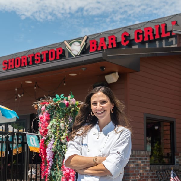 Smiling woman in chef's coat with arms crossed, standing outside Shortstop Bar & Grill.