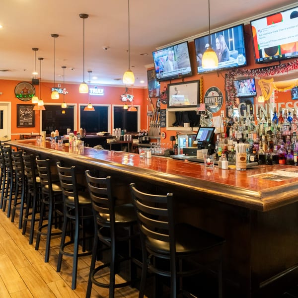 A long, well-stocked bar with numerous bottles, bar stools, and multiple TVs on the orange wall.