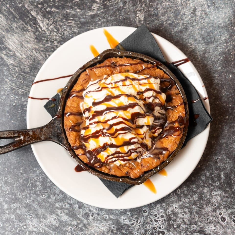 Skillet cookie with vanilla ice cream, chocolate, and caramel drizzle on a white plate.