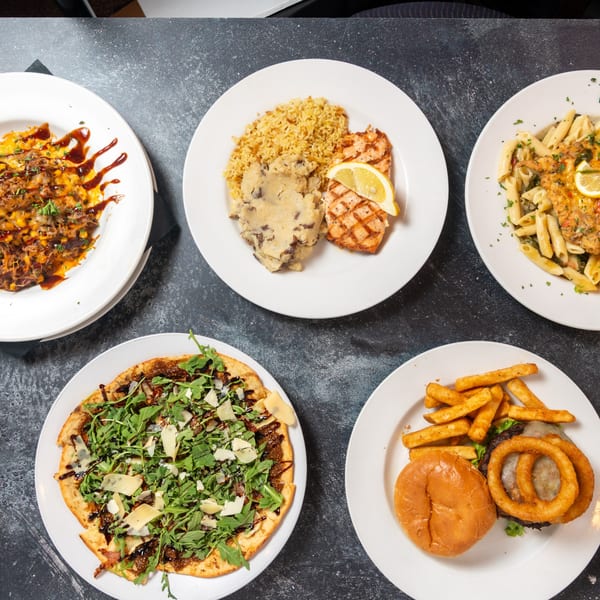 Overhead view of five plates of various restaurant dishes, including pasta, grilled salmon, and a burger.