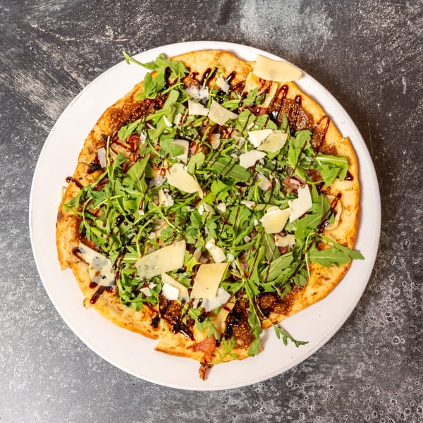 Flatbread with fig jam, prosciutto, arugula, parmesan, and balsamic glaze on a white plate.