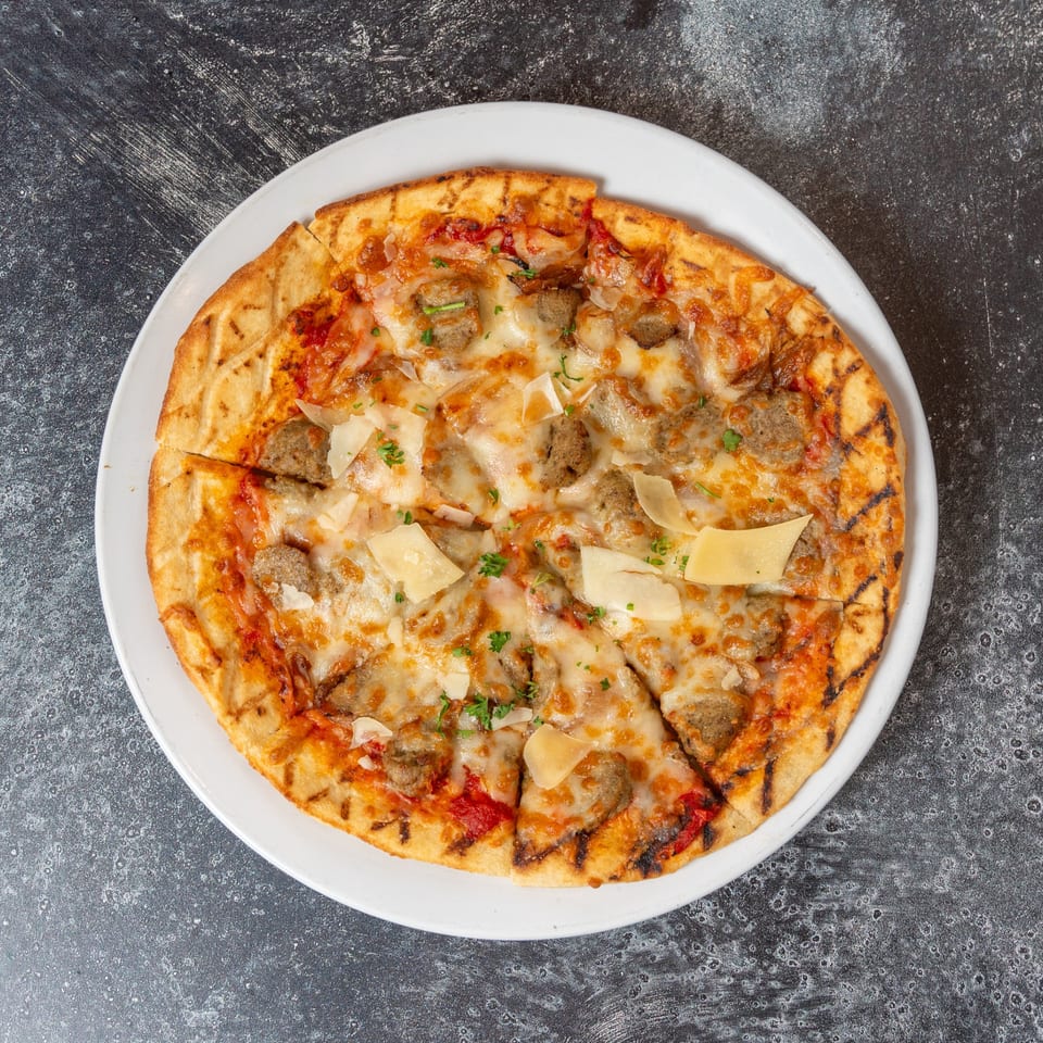 Grilled flatbread pizza with sausage, melted cheese, tomato sauce, and shaved parmesan on a white plate.