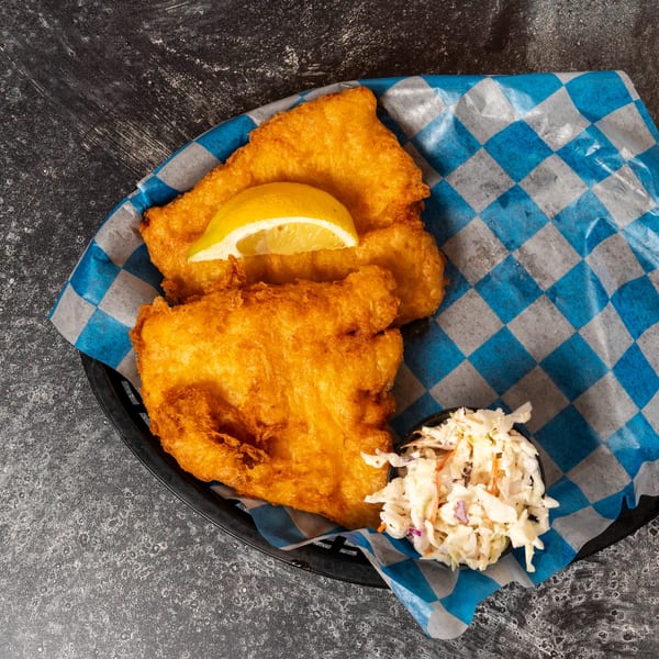 Golden-brown fried fish with a lemon wedge and a side of coleslaw in a basket with blue and white checkered paper.