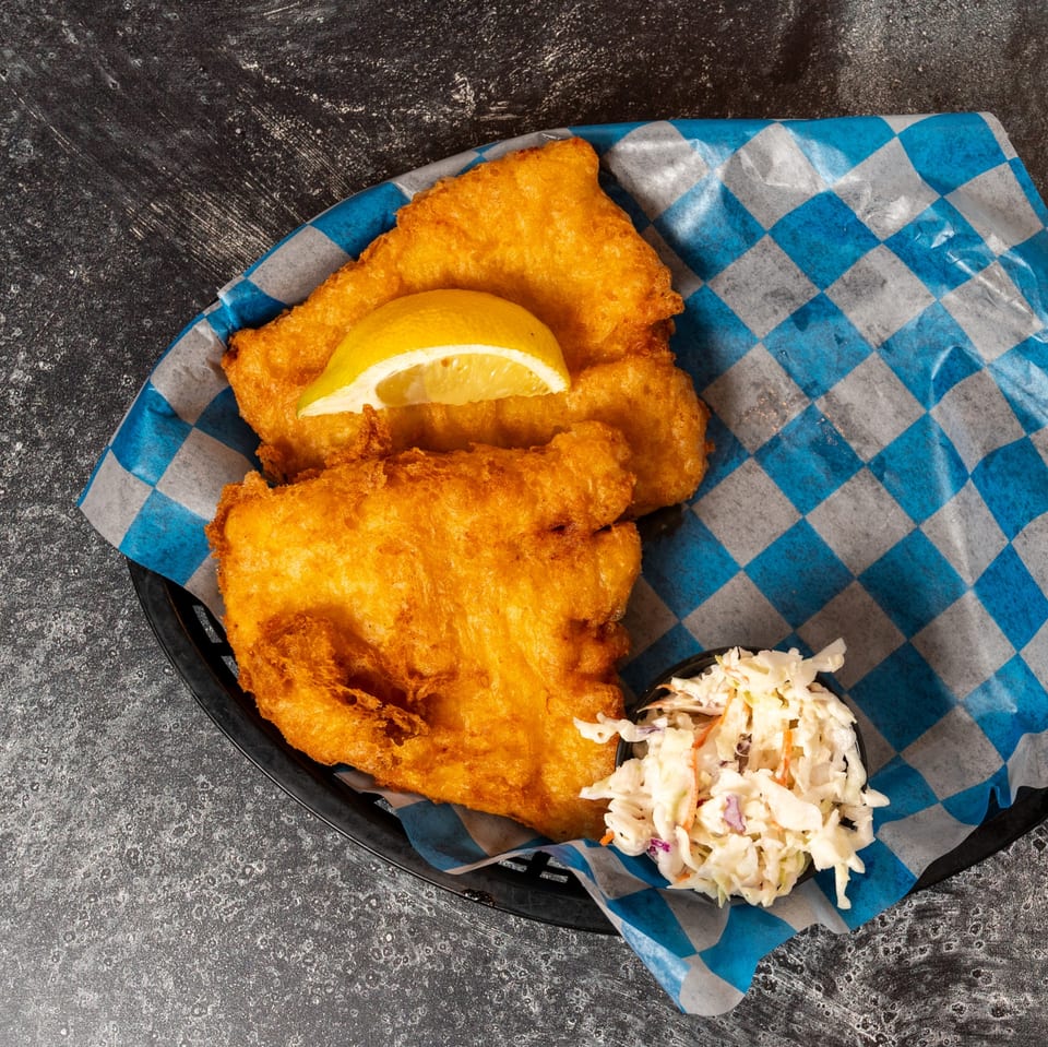 Golden-brown fried fish with a lemon wedge and a side of coleslaw in a basket with blue and white checkered paper.