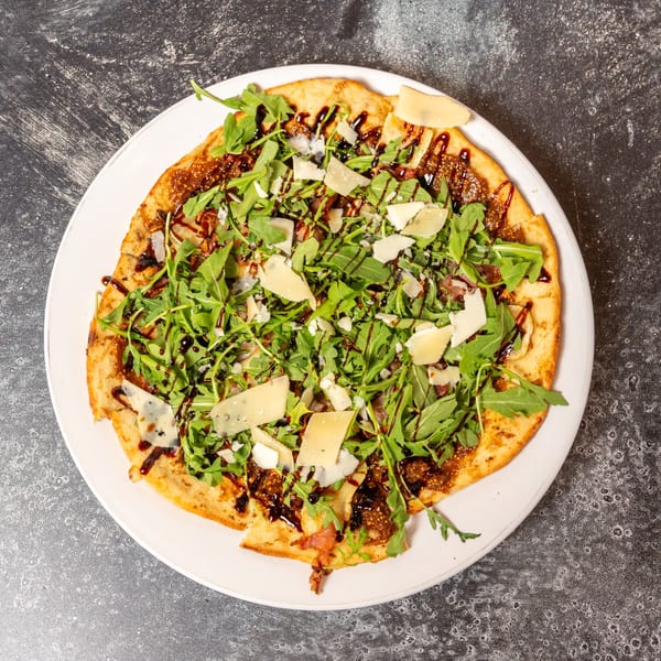Flatbread pizza topped with arugula, shaved Parmesan, fig jam, and balsamic glaze on a white plate.