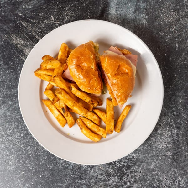 A white plate holds a sliced sandwich with ham and cheese, alongside seasoned potato wedges.