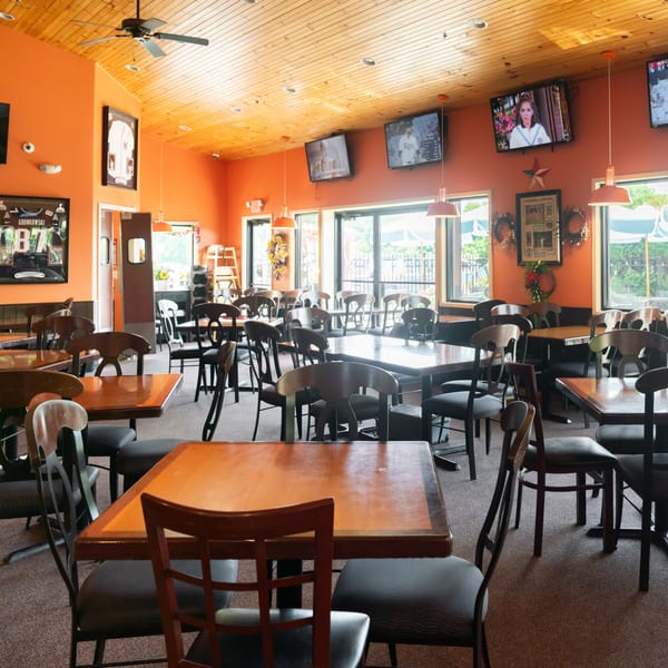 A restaurant dining room with orange walls, wooden tables, black chairs, and multiple TVs.